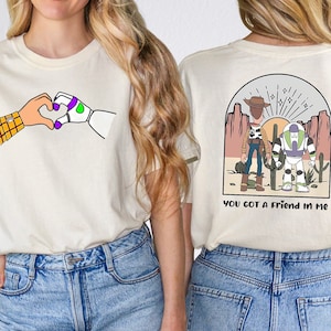 May include: Two white t-shirts with a Toy Story theme. One shirt features Woody and Buzz Lightyear holding hands with the text "You got a friend in me". The other shirt features Woody and Buzz Lightyear making a heart shape with their hands.
