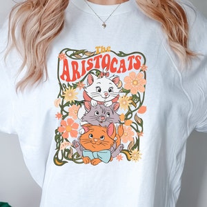May include: White t-shirt featuring a graphic of three cartoon cats from "The Aristocats" movie, surrounded by flowers and foliage. The text "The Aristocats" is displayed above the cats in a retro font. The cats are white, gray, and orange.