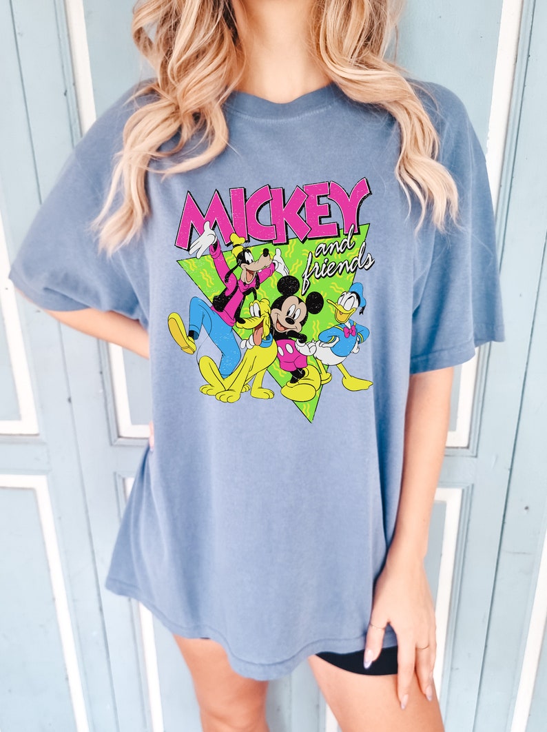 May include: A blue t-shirt with the text "MICKEY and friends" in pink and white, featuring cartoon characters Mickey Mouse, Goofy, Pluto, and Donald Duck. The shirt has short sleeves and a loose fit.