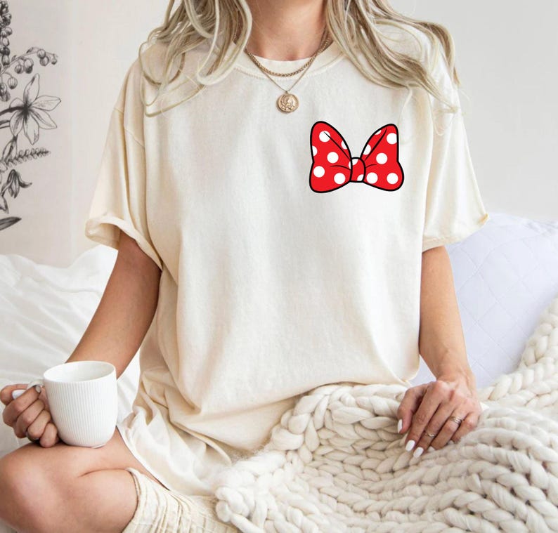 May include: Cream-colored t-shirt featuring a red bow with white polka dots on the chest. The bow has a black outline. The shirt has short sleeves and a relaxed fit. The person is wearing a gold necklace.