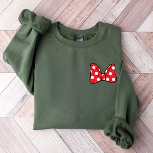 May include: An olive green sweatshirt featuring a red bow with white polka dots on the front. The sweatshirt has long sleeves with cuffed wrists and a crew neck. The bow is a classic design.