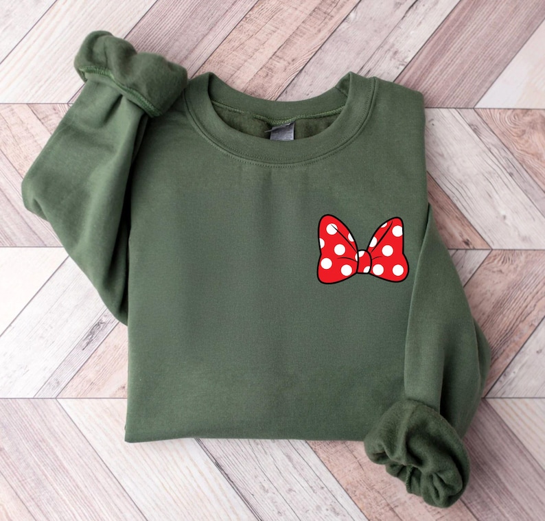 May include: An olive green sweatshirt featuring a red bow with white polka dots on the front. The sweatshirt has long sleeves with cuffed wrists and a crew neck. The bow is a classic design.