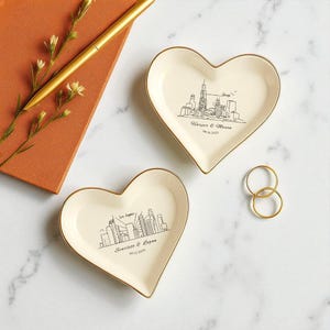 May include: Two heart-shaped trinket dishes with gold rims, featuring city skyline drawings. One dish displays "Chicago" and a date, the other "Los Angeles" and a date. A gold pen and a small notebook are in the background.