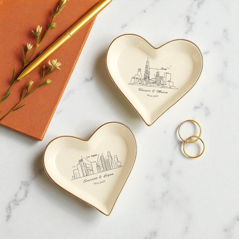 May include: Two heart-shaped trinket dishes with gold rims, featuring city skyline drawings. One dish displays "Chicago" and a date, the other "Los Angeles" and a date. A gold pen and a small notebook are in the background.