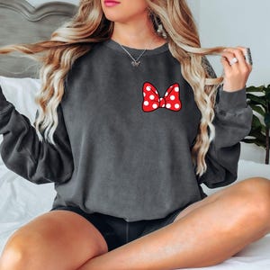 May include: Dark gray sweatshirt featuring a red bow with white polka dots. The bow is on the left chest area. The sweatshirt has long sleeves and a crew neck. The overall style is casual and comfortable.