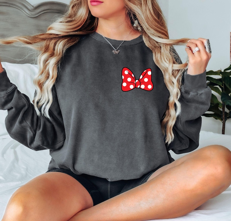 May include: Dark gray sweatshirt featuring a red bow with white polka dots. The bow is on the left chest area. The sweatshirt has long sleeves and a crew neck. The overall style is casual and comfortable.