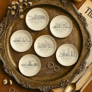 May include: A set of small, cream-coloured ceramic plates with gold edges, each displaying a black line drawing of a city skyline and names with dates. The plates are on a decorative tray with a candle and other items.