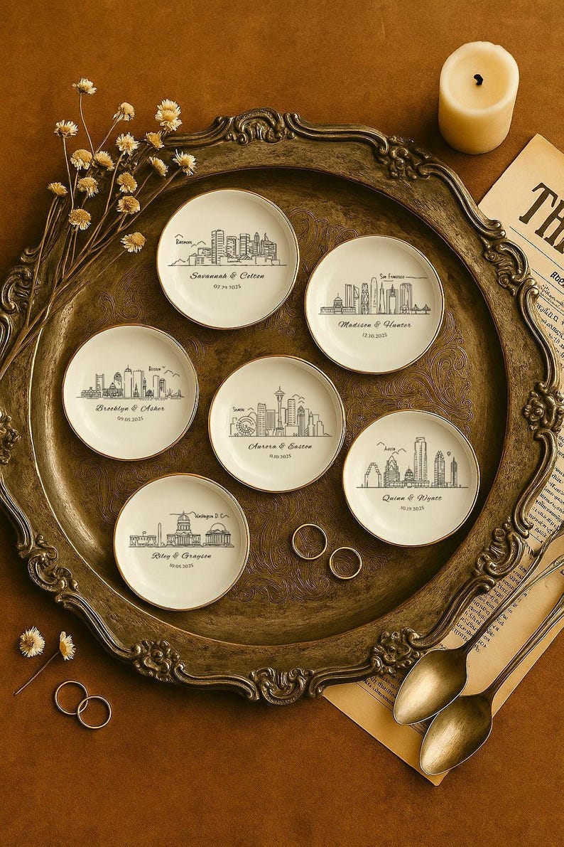 May include: A set of small, cream-coloured ceramic plates with gold edges, each displaying a black line drawing of a city skyline and names with dates. The plates are on a decorative tray with a candle and other items.