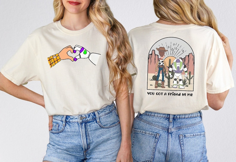 May include: Two white t-shirts with a Toy Story theme. One shirt features Woody and Buzz Lightyear holding hands with the text "You got a friend in me". The other shirt features Woody and Buzz Lightyear making a heart shape with their hands.