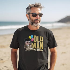 May include: Black t-shirt with a cartoon image of an older man with glasses wearing a Mickey Mouse hat and holding balloons. The text "OLD MAN COMING THROUGH" is printed in gold on the shirt.