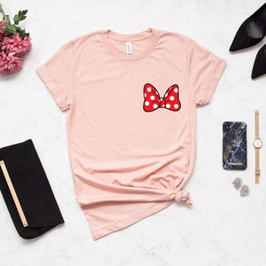 May include: A light pink t-shirt featuring a red bow with white polka dots on the chest. The bow is outlined in black. The shirt is styled with a knot at the bottom. Accessories include a black clutch, black heels, a marble phone case, a gold watch, and earrings.