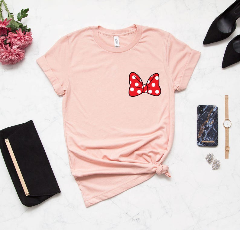 May include: A light pink t-shirt featuring a red bow with white polka dots on the chest. The bow is outlined in black. The shirt is styled with a knot at the bottom. Accessories include a black clutch, black heels, a marble phone case, a gold watch, and earrings.
