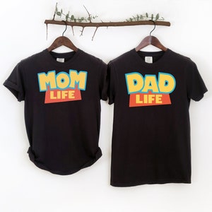 以下が含まれることがあります： トイストーリーをモチーフにしたデザインの黒いTシャツ2枚。左側のTシャツには「Mom Life」と黄色、青、赤で書かれています。右側のTシャツには「Dad Life」と黄色、青、赤で書かれています。