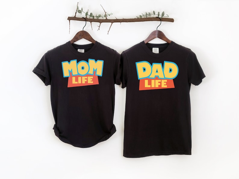以下が含まれることがあります： トイストーリーをモチーフにしたデザインの黒いTシャツ2枚。左側のTシャツには「Mom Life」と黄色、青、赤で書かれています。右側のTシャツには「Dad Life」と黄色、青、赤で書かれています。