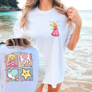 May include: White oversized t-shirt with a cartoon image of Princess Peach from the Super Mario Bros. video game. The text "Princess Peach" is printed above the image. The back of the shirt features a light blue background with four cartoon images: Princess Peach, Toad, Yoshi, and a star.