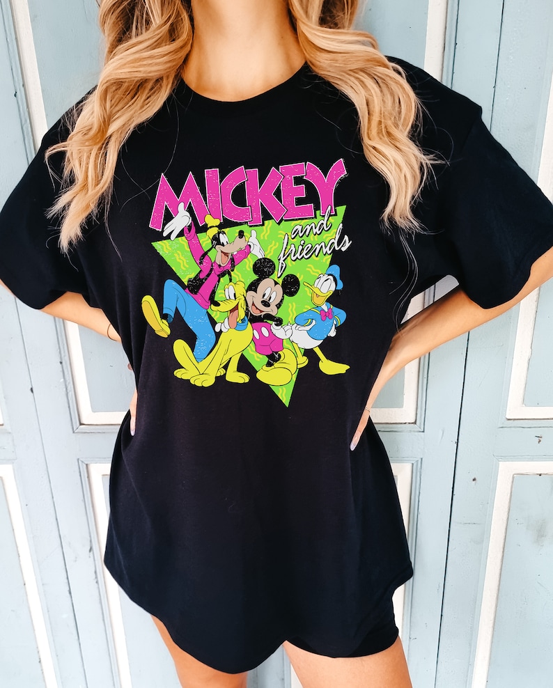 May include: Black t-shirt with a colourful graphic of Mickey Mouse and friends. The design features Mickey, Donald Duck, Goofy, and Pluto, with the words "Mickey and friends" in pink and white. The characters are set against a green triangle.