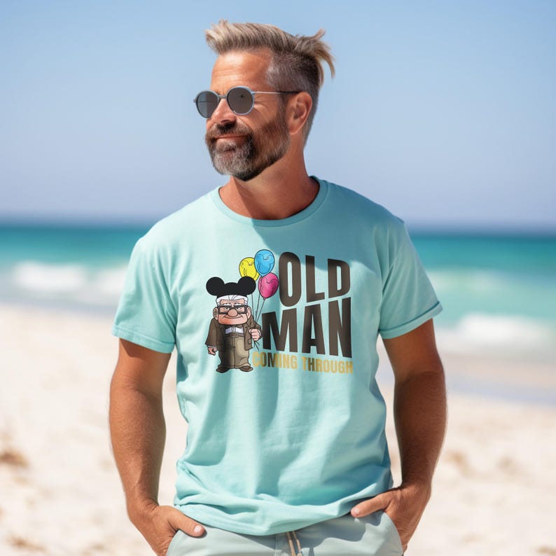 May include: A light blue t-shirt with a cartoon illustration of an older man with a mustache and glasses holding balloons. The text "OLD MAN COMING THROUGH" is printed in gold on the shirt.