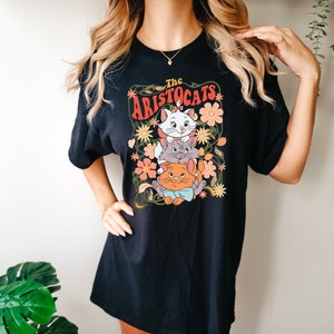 May include: Black t-shirt featuring a colorful graphic of three cartoon cats from "The Aristocats" movie, surrounded by flowers and vines. The text "The Aristocats" is arched above the cats in orange and white lettering. The shirt is a casual, oversized style.