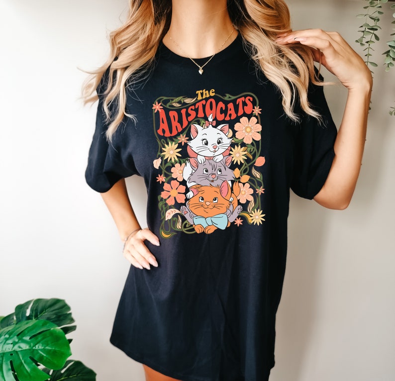 May include: Black t-shirt featuring a colorful graphic of three cartoon cats from "The Aristocats" movie, surrounded by flowers and vines. The text "The Aristocats" is arched above the cats in orange and white lettering. The shirt is a casual, oversized style.