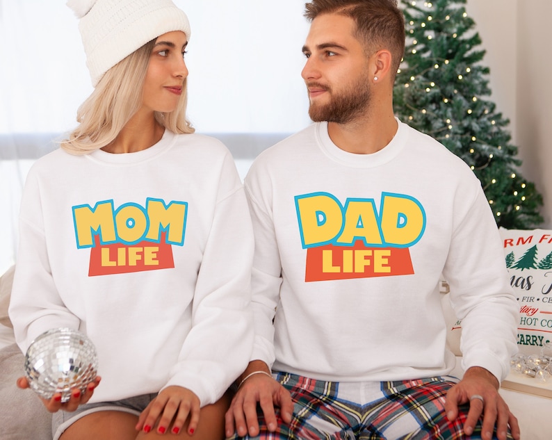 以下が含まれることがあります： カラフルな漫画風のフォントで「Mom Life」と「Dad Life」と書かれた白いスウェットシャツ。スウェットシャツはセットになっています。