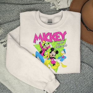 May include: A cream-coloured sweatshirt with a graphic of Mickey Mouse and friends. The design features Mickey, Goofy, Donald Duck, and Pluto, with the words "Mickey and Friends" in pink and green. The sweatshirt is folded on a patterned surface.