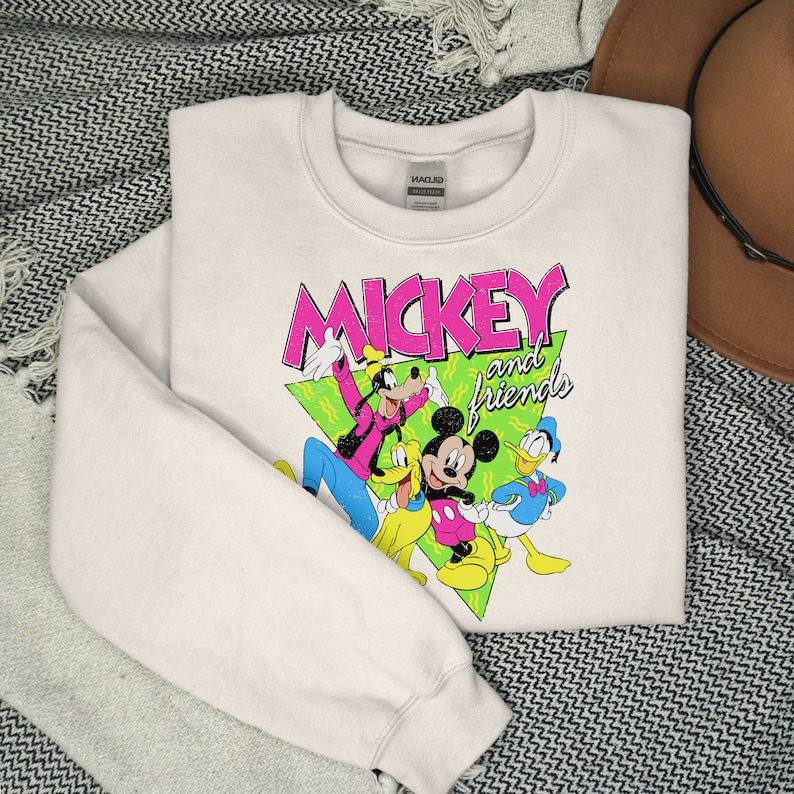 May include: A cream-coloured sweatshirt with a graphic of Mickey Mouse and friends. The design features Mickey, Goofy, Donald Duck, and Pluto, with the words "Mickey and Friends" in pink and green. The sweatshirt is folded on a patterned surface.