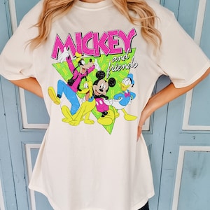 May include: Cream-coloured t-shirt with the text "MICKEY and friends" in pink and white, featuring cartoon characters Mickey Mouse, Goofy, Pluto, and Donald Duck. The graphic is set against a green triangle.