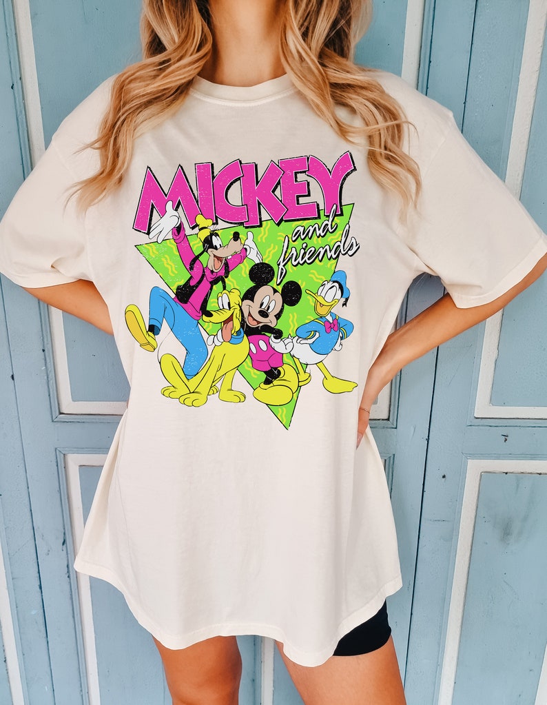 May include: Cream-coloured t-shirt with the text "MICKEY and friends" in pink and white, featuring cartoon characters Mickey Mouse, Goofy, Pluto, and Donald Duck. The graphic is set against a green triangle.