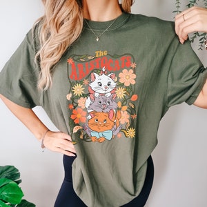 May include: Olive green t-shirt featuring a graphic of three cartoon cats from "The Aristocats" with floral accents. The text "The Aristocats" is in red. The shirt has short sleeves and a crew neck.