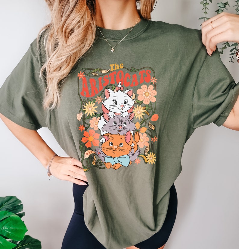 May include: Olive green t-shirt featuring a graphic of three cartoon cats from "The Aristocats" with floral accents. The text "The Aristocats" is in red. The shirt has short sleeves and a crew neck.