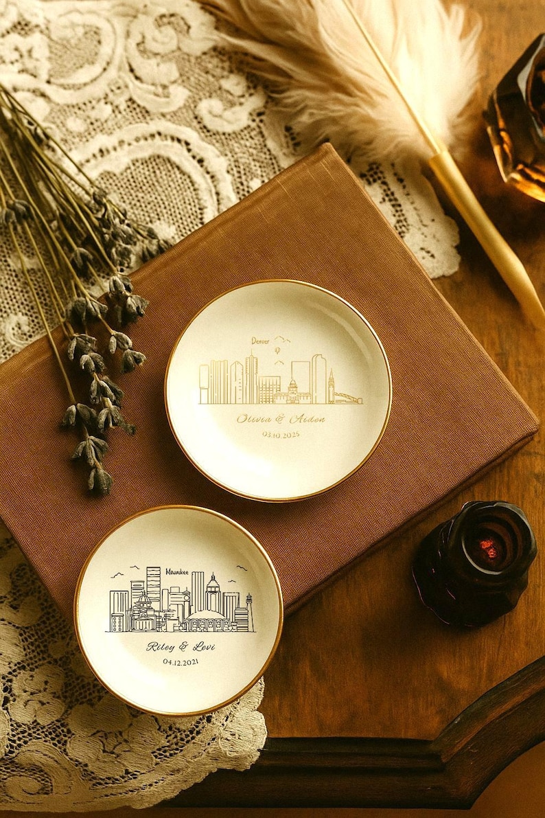 May include: Two small, white ceramic dishes with gold rims, each featuring a city skyline design. One dish is inscribed with "Denver" and the names "Olivia & Aiden" and a date. The other dish says "Milwaukee" and the names "Riley & Levi" and a date.