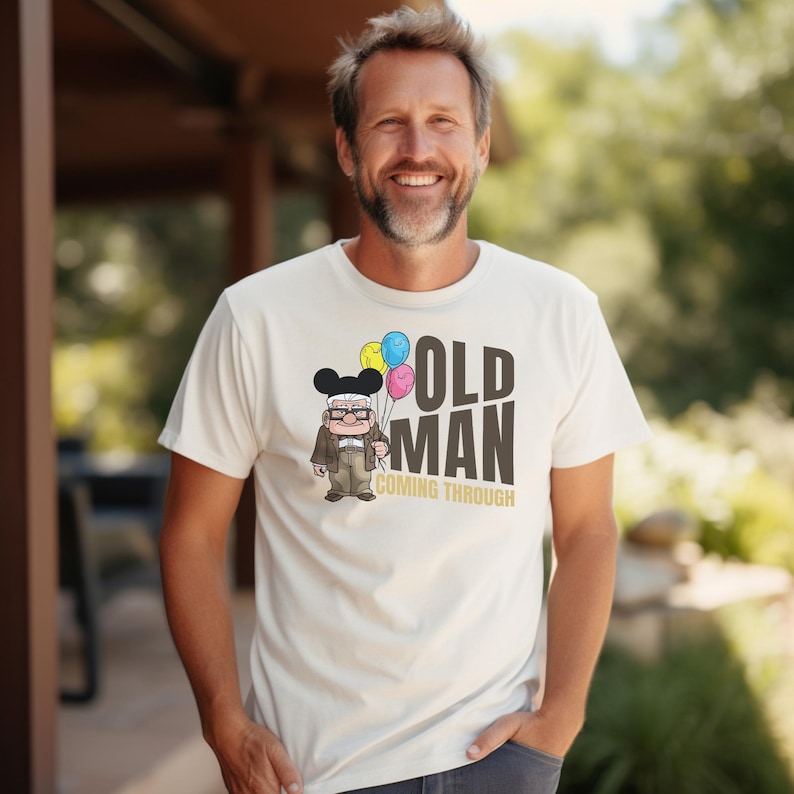 May include: White t-shirt with a cartoon illustration of an older man with glasses wearing a Mickey Mouse hat and holding balloons. The text "OLD MAN COMING THROUGH" is printed on the shirt.