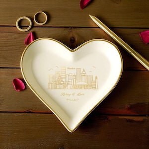 May include: A heart-shaped, cream-coloured ceramic trinket dish with a gold rim and a gold skyline design. The dish has the words "Riley & Love" and the date 04.12.2021. Two gold rings and a gold pen are also visible.