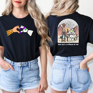 May include: Two black t-shirts with a Toy Story theme. The front shirt features Woody and Buzz Lightyear holding hands in a heart shape. The back shirt features Woody and Buzz Lightyear standing in a desert landscape with the text "You got a friend in me".