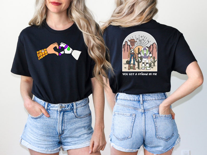 May include: Two black t-shirts with a Toy Story theme. The front shirt features Woody and Buzz Lightyear holding hands in a heart shape. The back shirt features Woody and Buzz Lightyear standing in a desert landscape with the text "You got a friend in me".