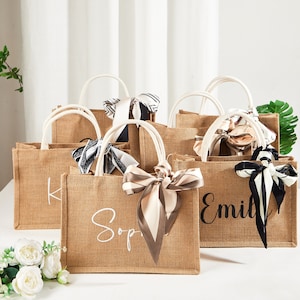 Könnte beinhalten: Mehrere Tragetaschen aus Jute mit weißen Griffen und dekorativen Seidenschals. Die Taschen tragen Namen in weißer Schrift wie "Sophie" und "Emily". Ideal für Geschenke, eignen sie sich für Hochzeiten oder Partys.