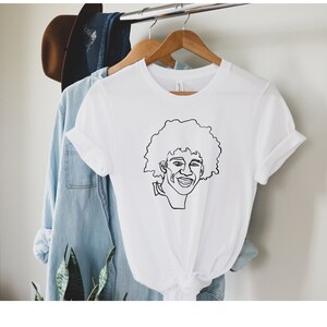 May include: White t-shirt featuring a black line drawing of a person with curly hair smiling.