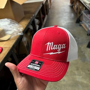 May include: Red and white trucker hat with a white embroidered lightning bolt and the word "Maga" on the front. The hat is made by Richardson 112.