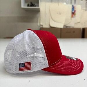 May include: A red and white trucker hat with a mesh back. The hat has a small American flag patch on the front.