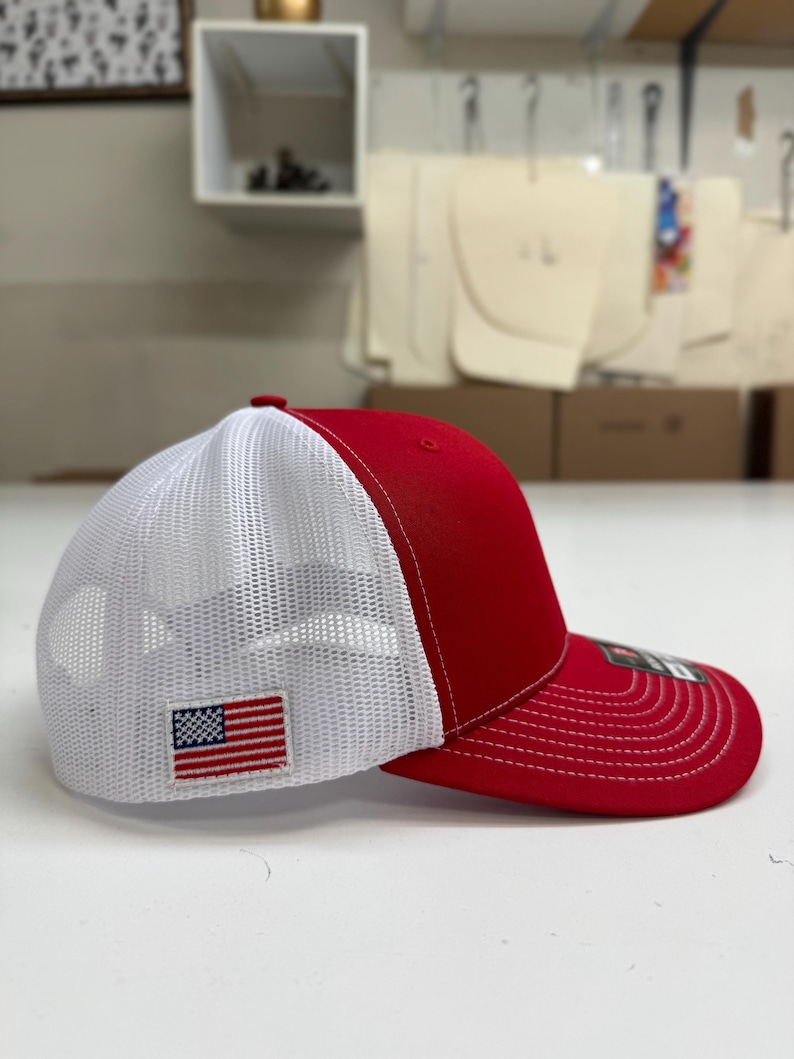 May include: A red and white trucker hat with a mesh back. The hat has a small American flag patch on the front.