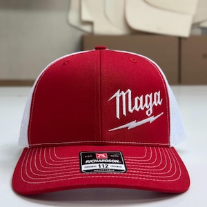 May include: A red and white trucker hat with a white embroidered logo that says "Maga" with a lightning bolt. The hat is adjustable and has a black label that says "Richardson 112 Original Trucker Adjustable".
