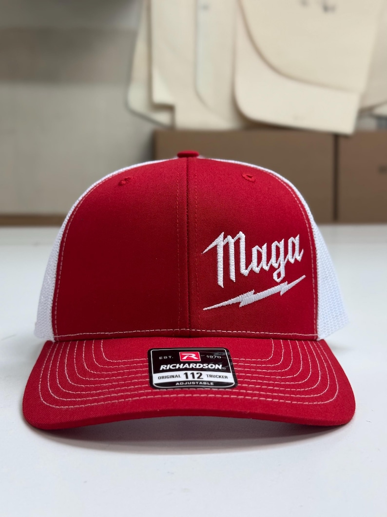 May include: A red and white trucker hat with a white embroidered logo that says "Maga" with a lightning bolt. The hat is adjustable and has a black label that says "Richardson 112 Original Trucker Adjustable".