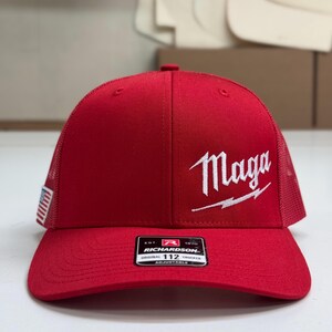 May include: Red trucker hat with a white "Maga" logo and a lightning bolt. The hat has a black and white American flag patch on the side and a black and white "Richardson" label on the back.