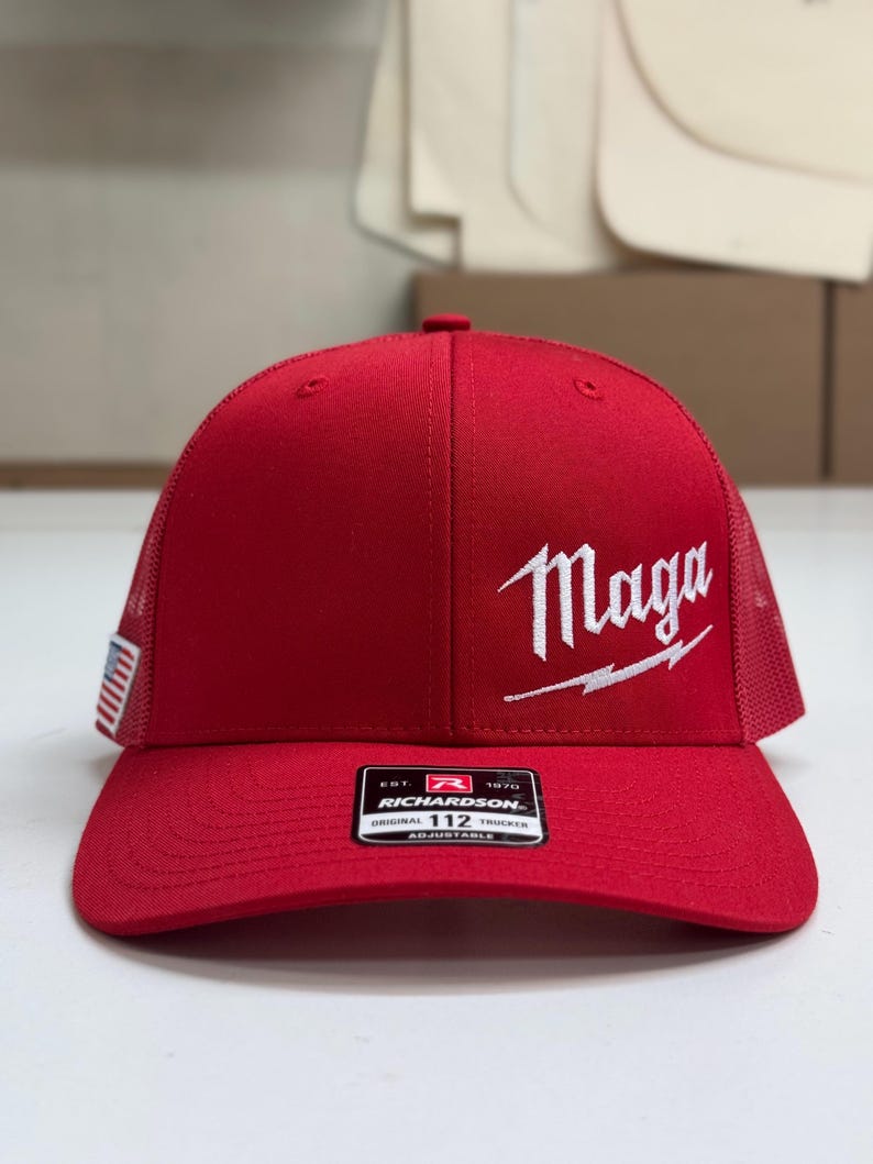 May include: Red trucker hat with a white "Maga" logo and a lightning bolt. The hat has a black and white American flag patch on the side and a black and white "Richardson" label on the back.