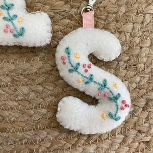 May include: White felt letter "S" with teal, pink, and yellow floral embroidery. It has a pink ribbon and silver clasp. The background is a woven, tan material.