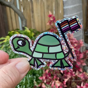 May include: A green turtle with a sparkly iridescent finish holding a pride flag with pink, white, and light blue stripes. The flag is attached to a stick.