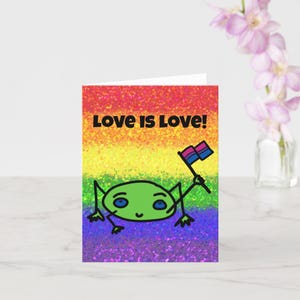 May include: A greeting card with a rainbow glitter background and a green frog holding a bisexual pride flag. The text "Love is Love!" is printed in black at the top of the card.