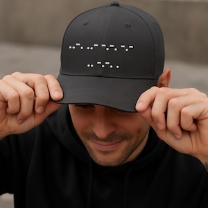 May include: A charcoal grey baseball cap featuring white Morse code symbols. The cap has a classic six-panel construction and a curved peak. The Morse code likely spells out a word or phrase.