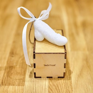Puede incluir: Adorno blanco brillante con forma de mano con un lazo de cinta blanca. El adorno está sentado en una pequeña caja de madera con el texto 'Jack Frost' grabado en la parte delantera.