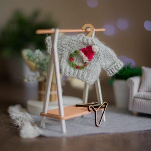 Op de afbeelding: Een miniatuur grijze trui met een kerstkrans en kerstmuts ontwerp hangt aan een houten hanger. De trui wordt getoond op een kleine witte en houten kledingrek. Een miniatuur kledingrek en een kleine bank staan op de achtergrond.
